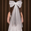 A white bridal veil with a large matching bow on the back, worn by a model shown from behind against a brown curtain background.