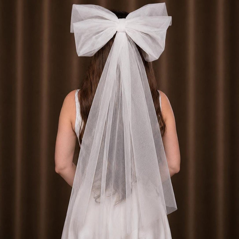 A white bridal veil with a large matching bow on the back, worn by a model shown from behind against a brown curtain background.